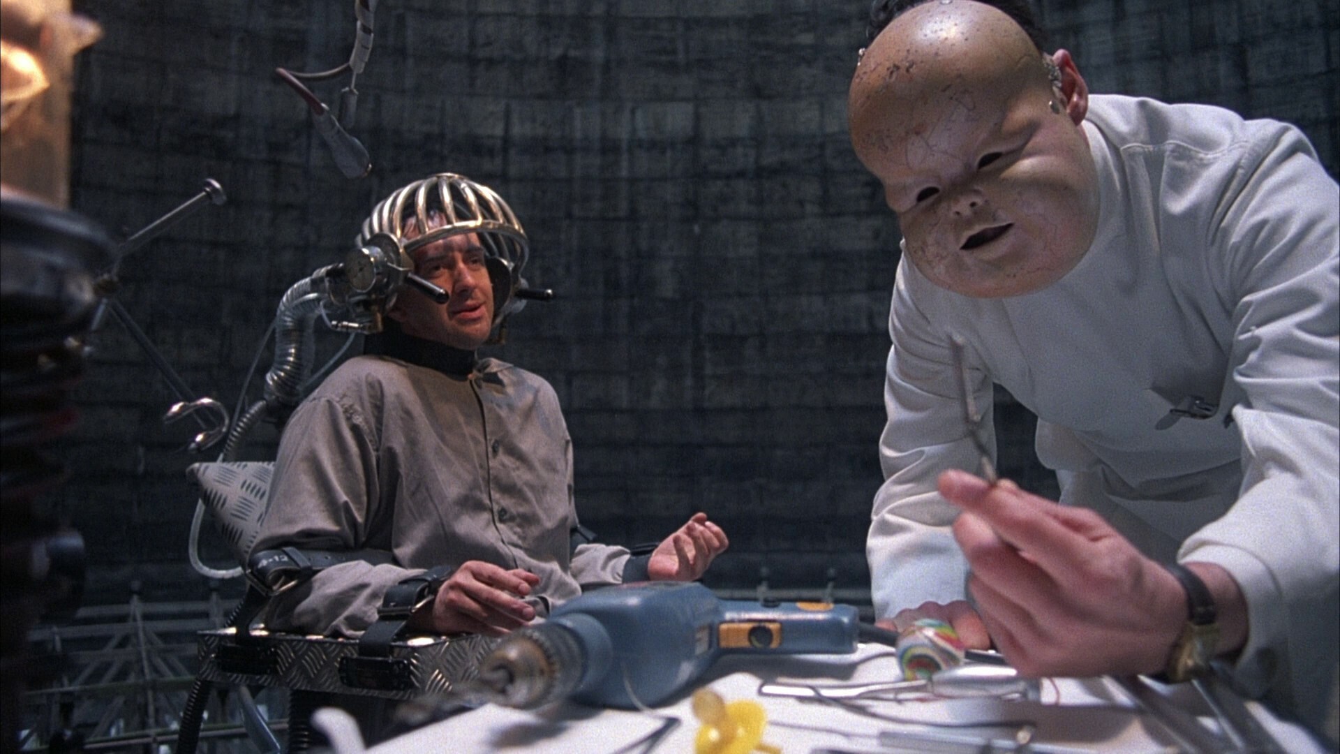 Le héros en mauvaise posture dans une chaise avec un tortionnaire portant un masque de bébé 