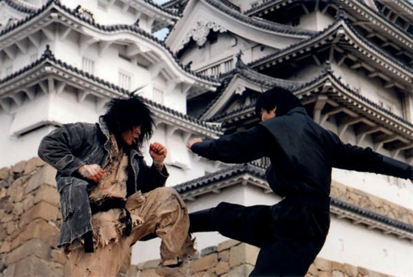 Choi Bae-Dal qui bloque en mawashi avec sa jambe gauche. Il est en tenue usée et le mec en face en tenue de ninja noir. Le temple derrière contient plusieurs toitures typiques avec des murs blancs